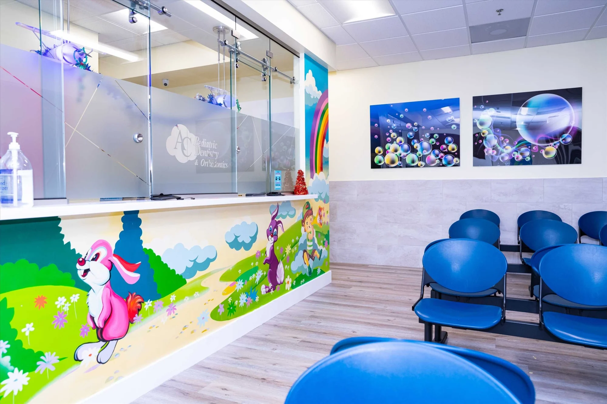 AC Pediatric Dentistry & Orthodontics — Reception counter with a mural of rabbits and a rainbow, facing rows of blue waiting chairs