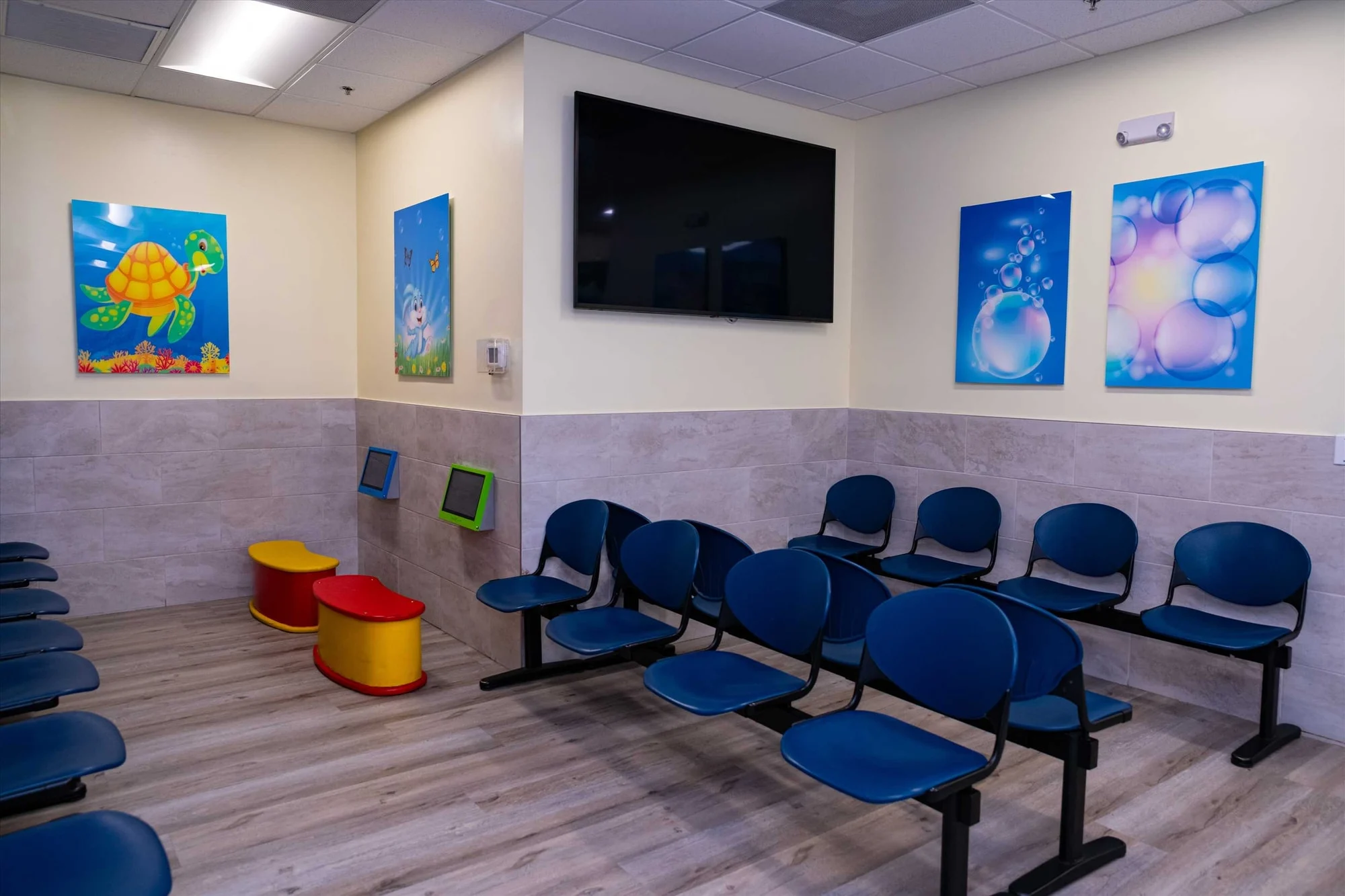 AC Pediatric Dentistry & Orthodontics — Waiting room with rows of blue chairs, a wall-mounted TV and red-and-yellow children's seats