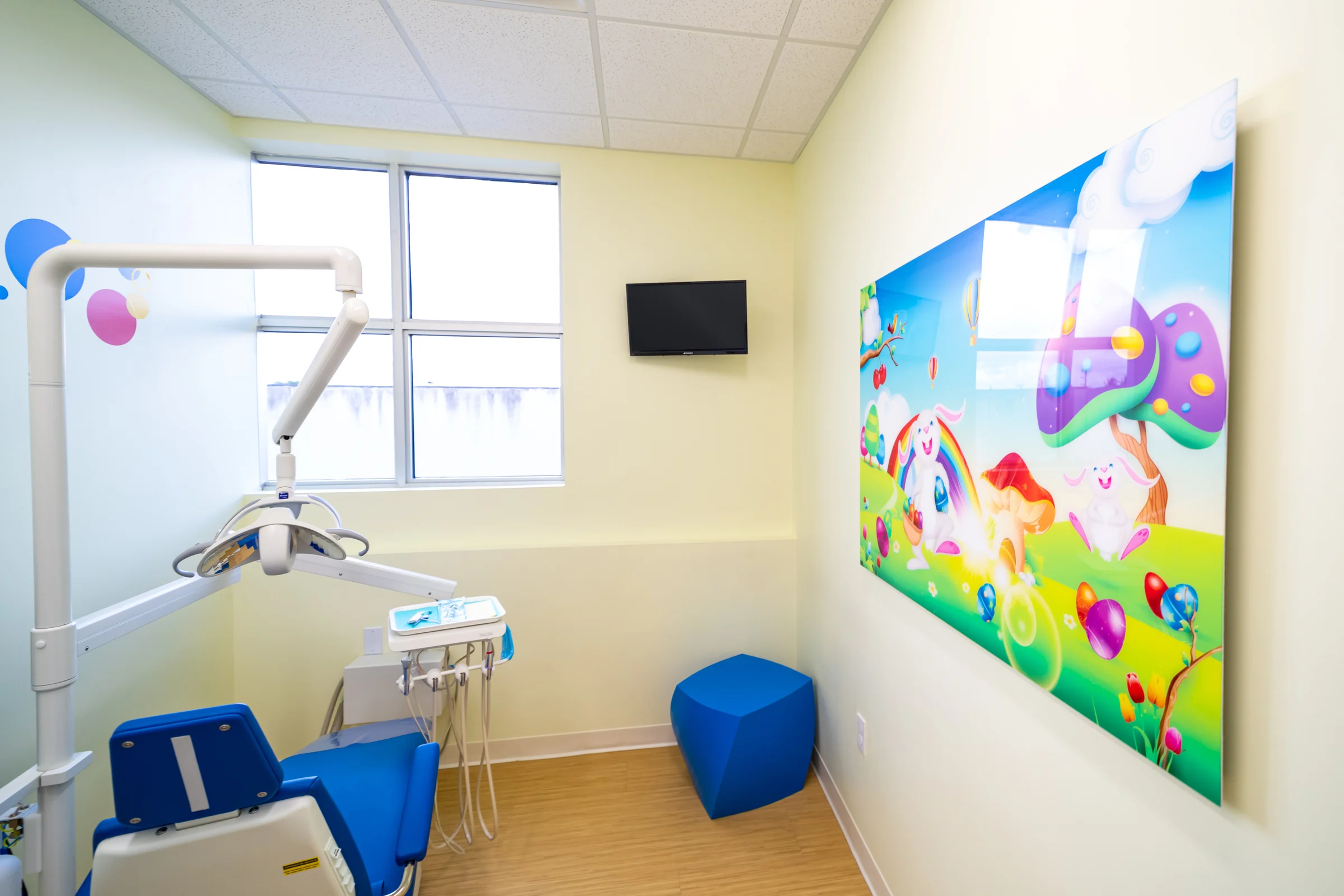 AC Pediatric Dentistry & Orthodontics — Treatment room with a large art panel of bunnies and a rainbow, a blue stool and a window behind the dental chair