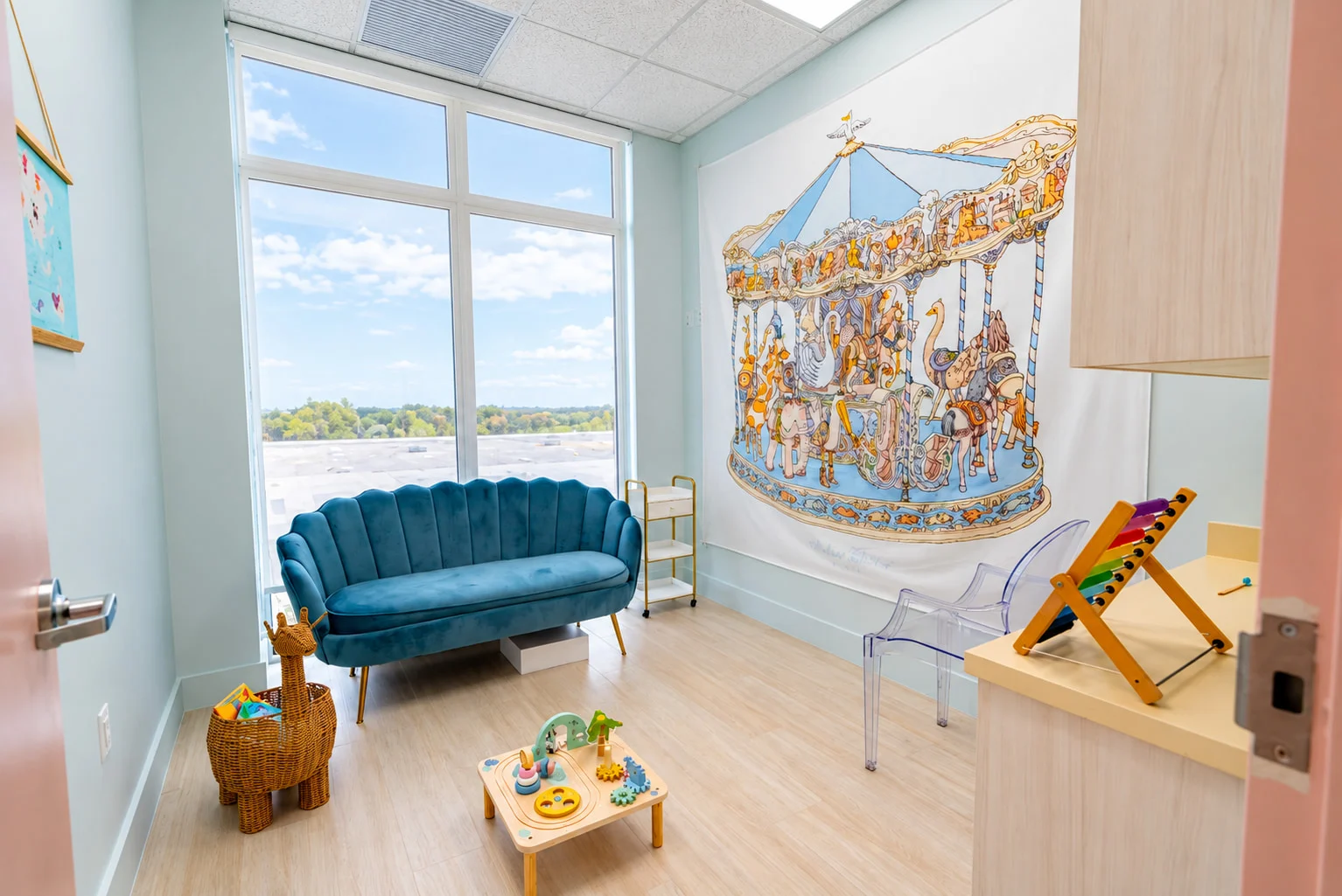 Denti Pediatric Dentistry — Small playroom with a teal velvet loveseat, a large carousel wall hanging and a floor-to-ceiling window