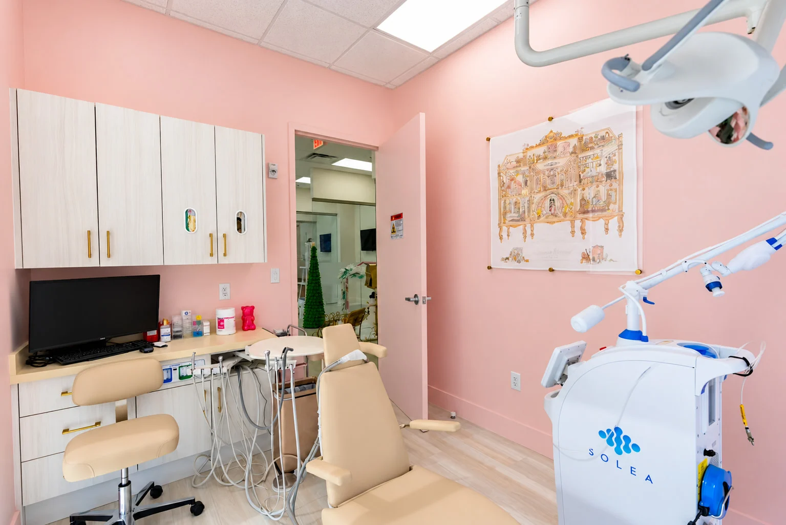 Denti Pediatric Dentistry — Treatment room with pink walls, a beige dental chair and light wood-look cabinets with brass-toned pulls