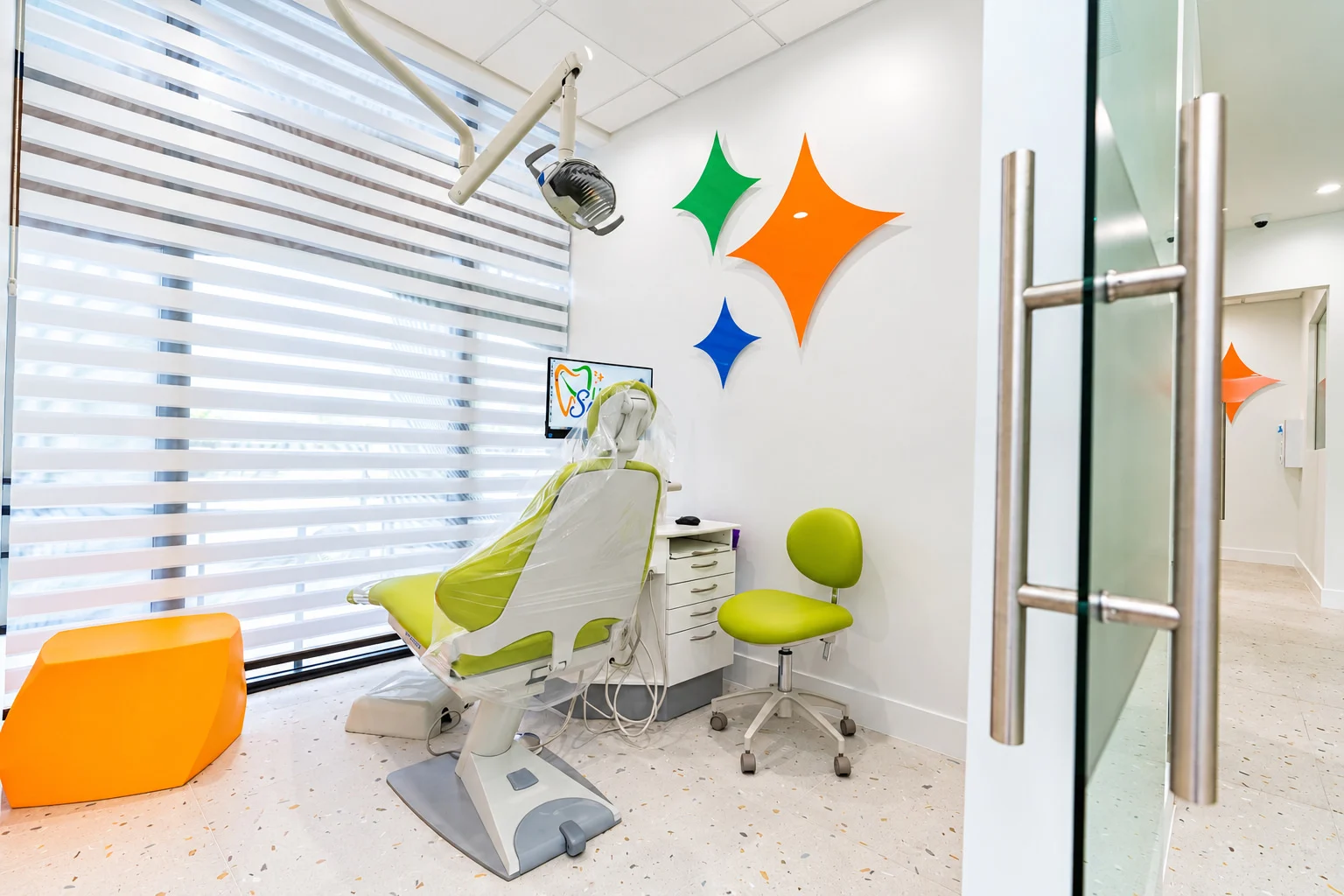Little Sparkle Pediatric Dentistry — Treatment room with a lime-green dental chair and stool, and three four-pointed stars in orange, green and blue on the wall