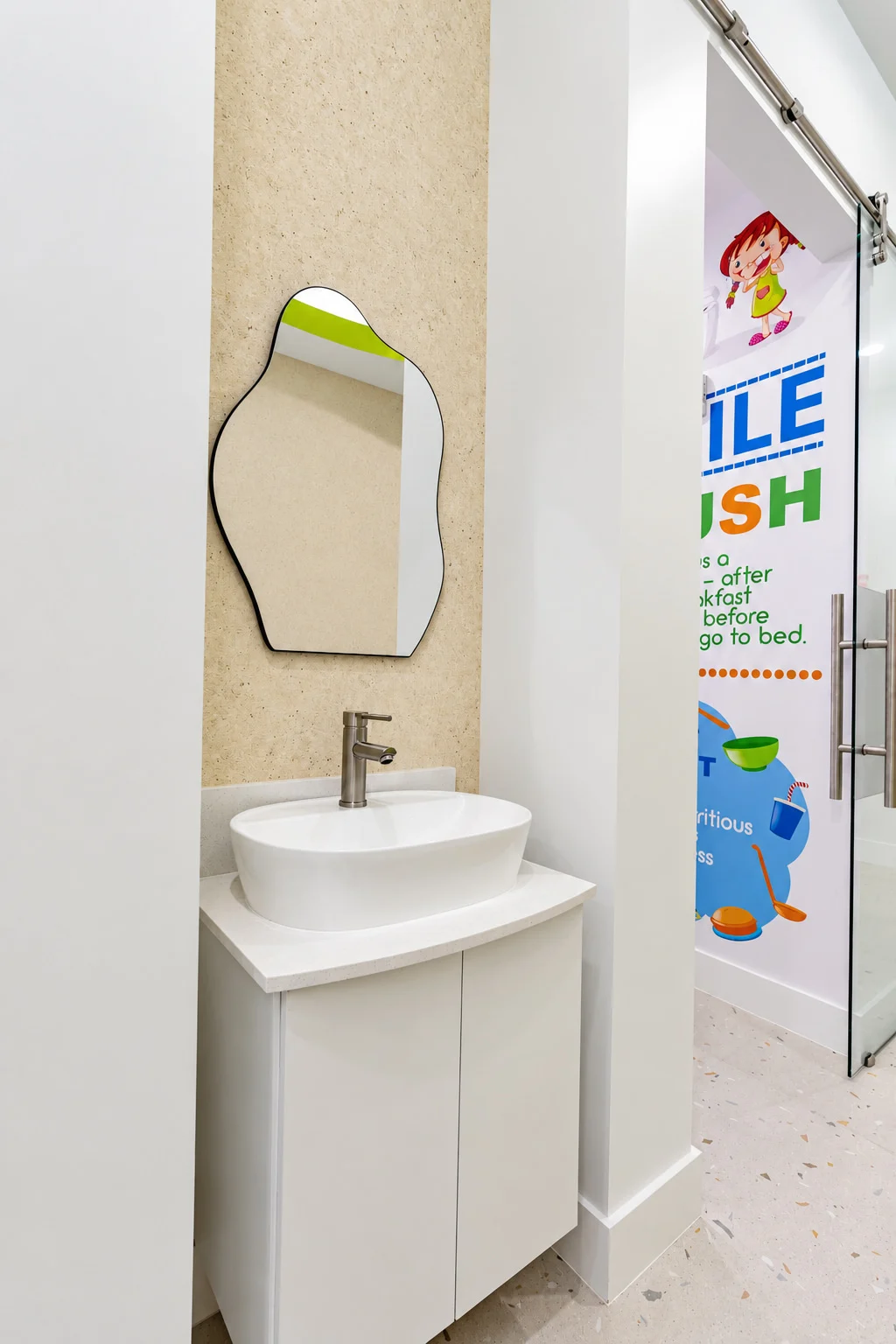 Little Sparkle Pediatric Dentistry — Bathroom nook with a white vessel basin under an organic-shaped mirror, next to a glass door printed with brushing advice
