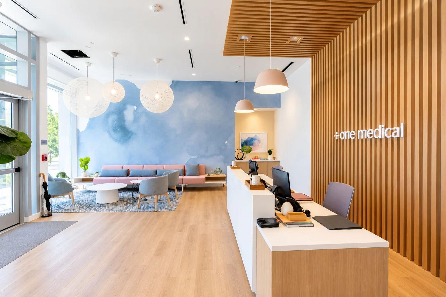 One Medical — Reception desk beside a timber-slat wall with a lettered sign, facing pink seating under a blue watercolor wall