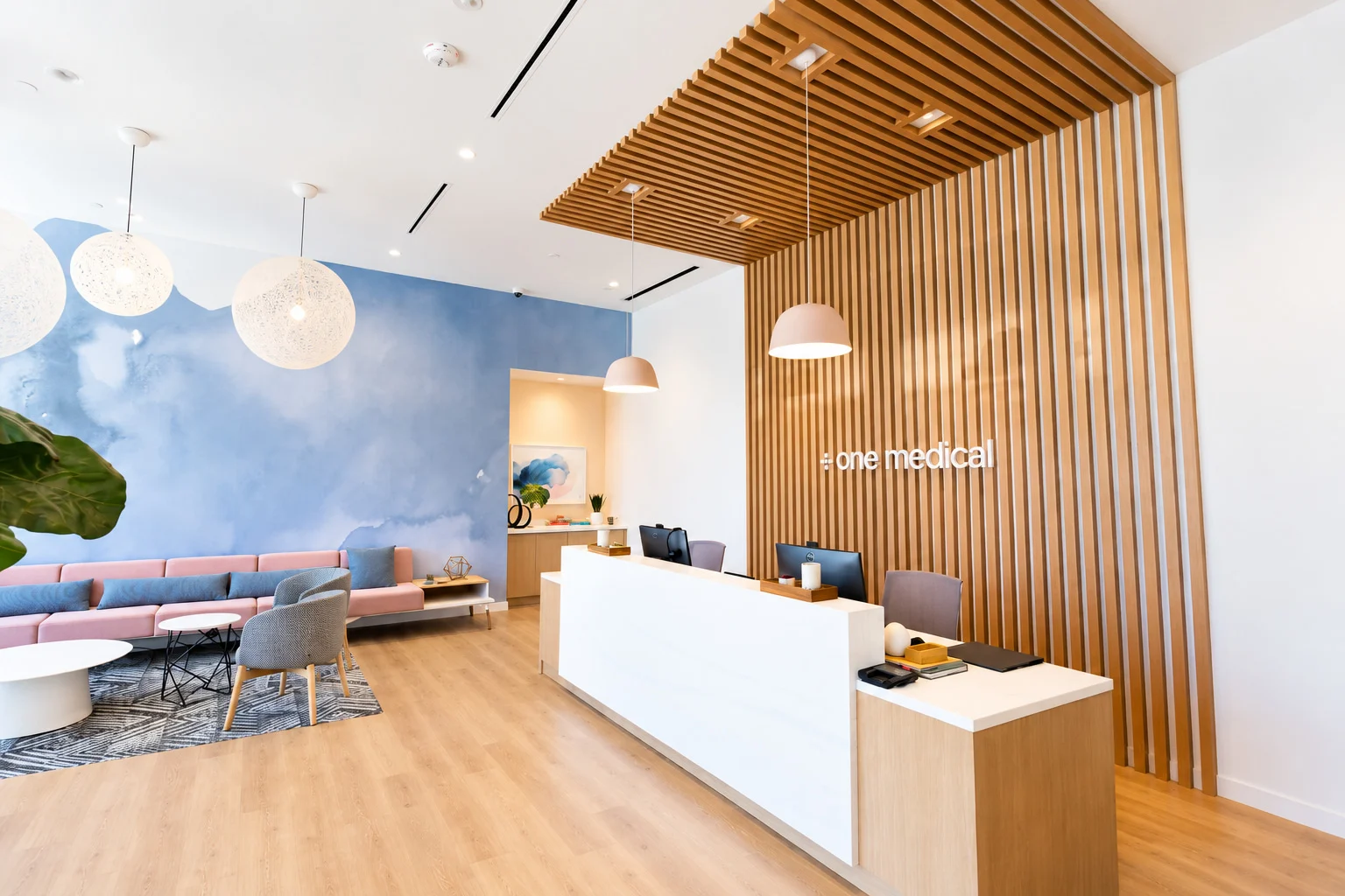 One Medical — Reception under a timber-slat canopy with pink dome pendants, and behind it a blue watercolor wall with woven globe lamps