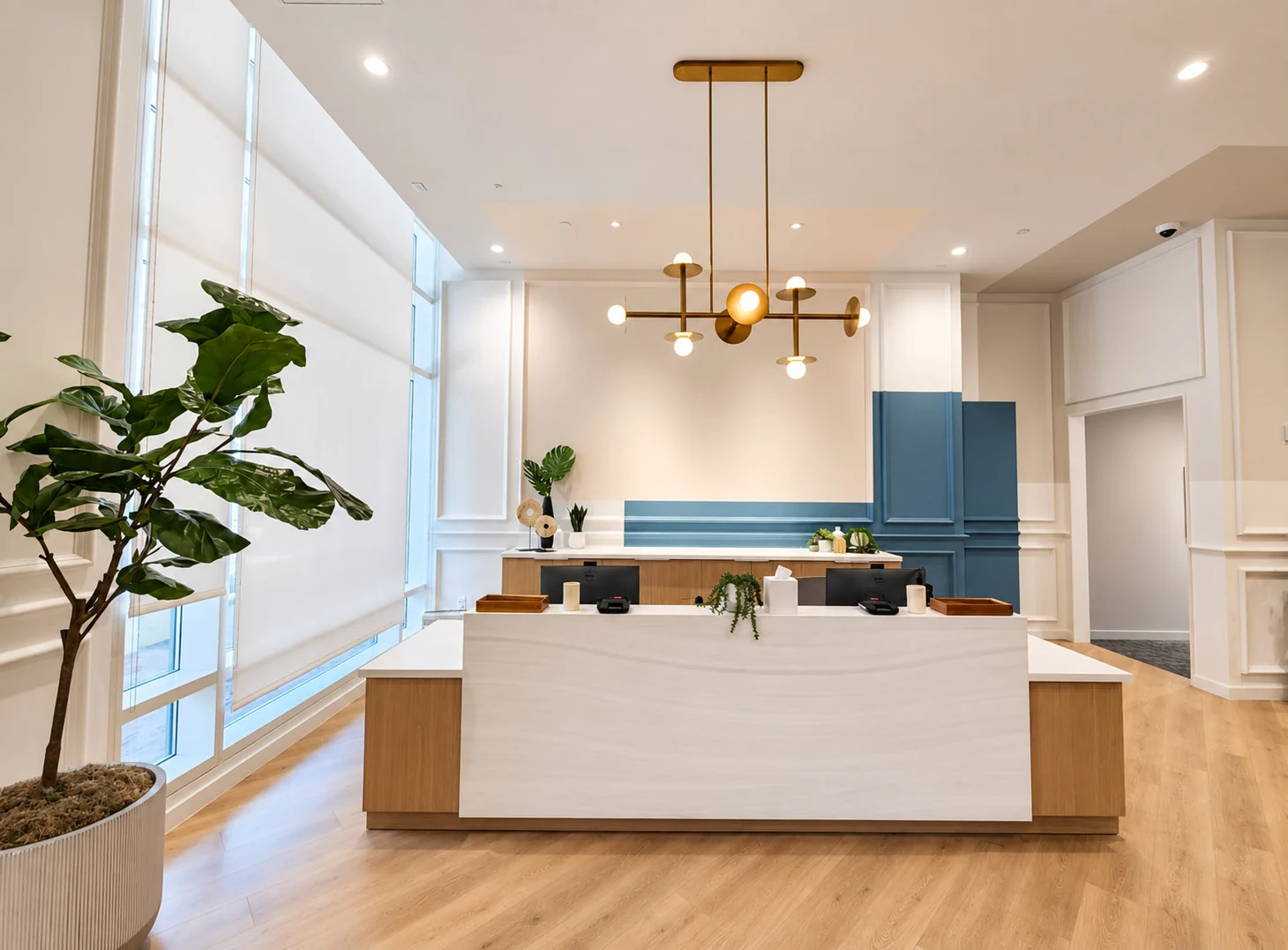 One Medical — Pale reception desk with timber sides under a brass chandelier, with white wall paneling and a block of blue