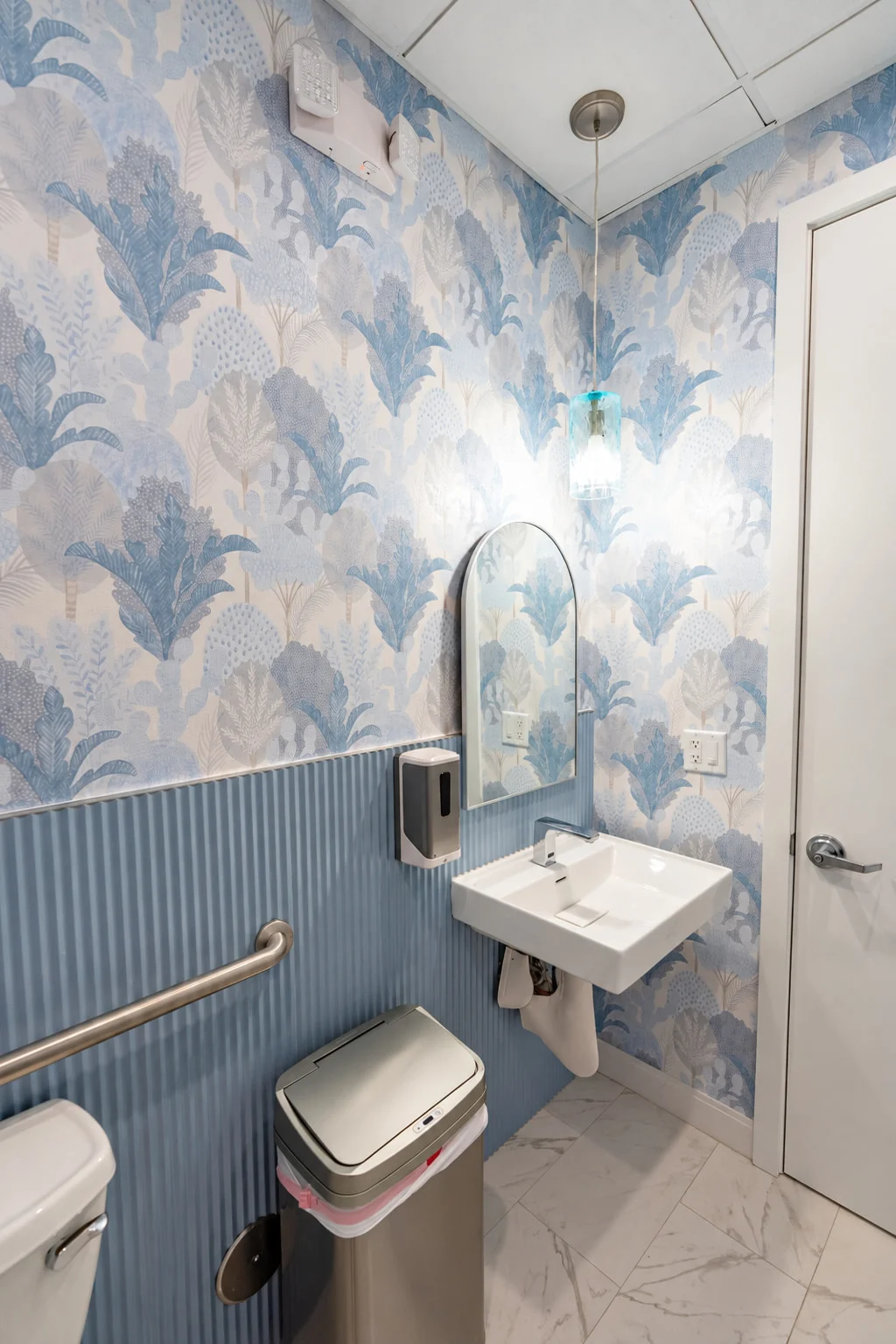 Specialty Smiles Orthodontics — Restroom with blue botanical wallpaper above ribbed blue wainscoting, an arched mirror and a wall-hung sink